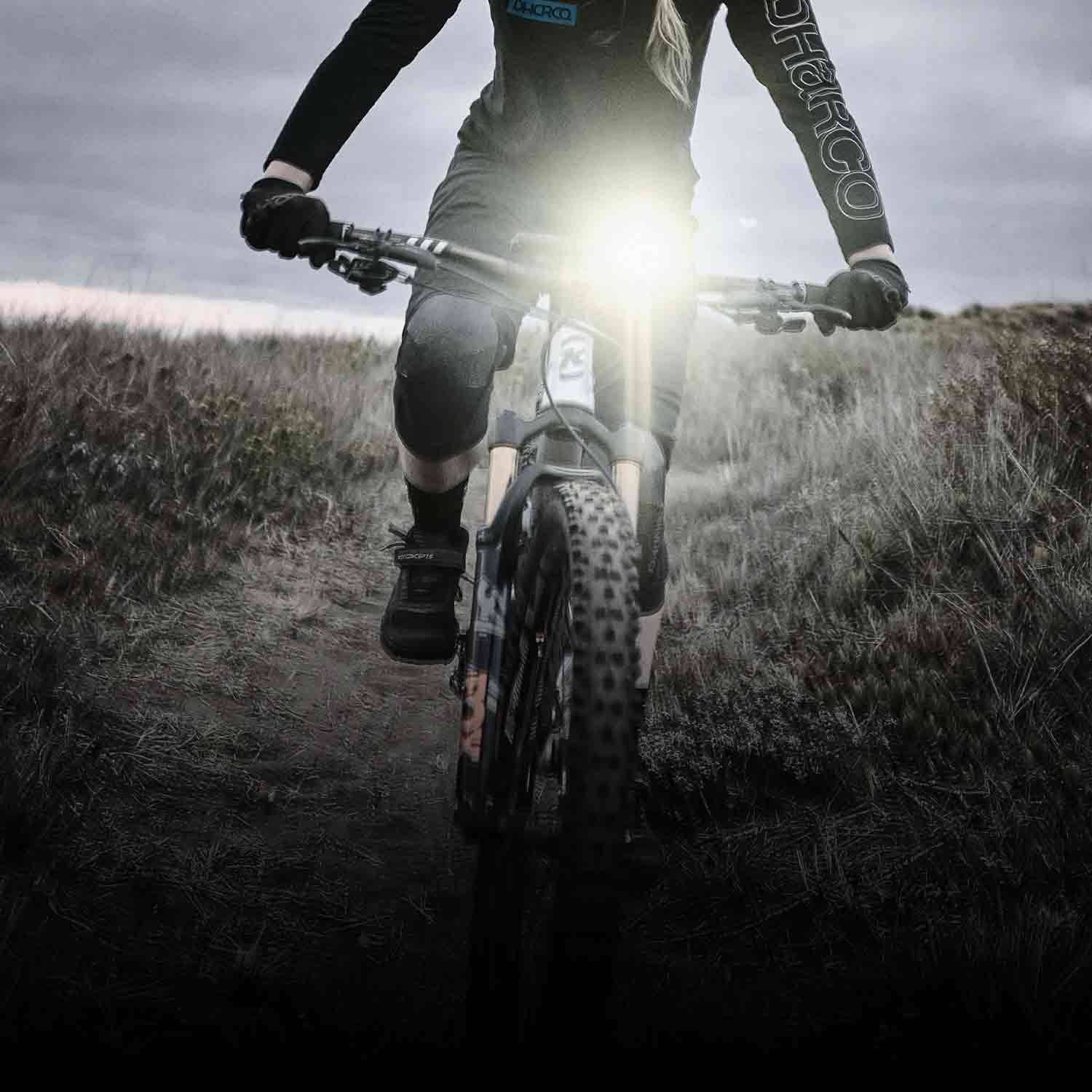 A bright front light on a bike being ridden on a trail.