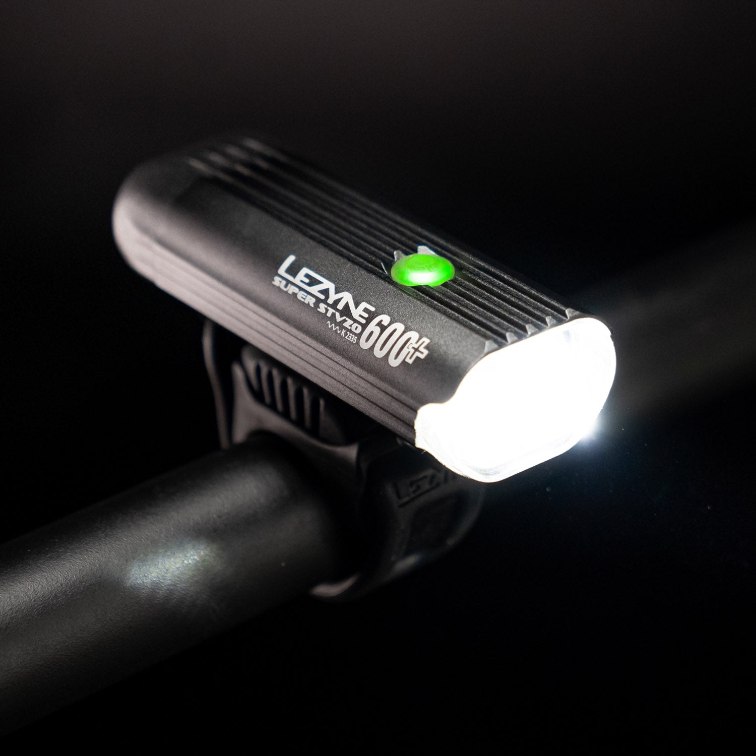 Top-down side view of the Lezyne Super StVZO 600+ LED frontlight, with a green circular power light on top, and emitting a bright white beam, mounted on the handlebars.