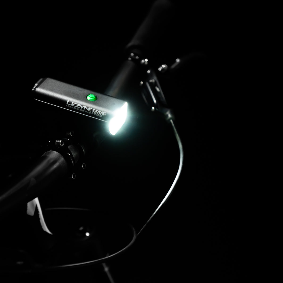 Top view of MACRO DRIVE 1400+ front LED bike light, mounted on gravel bike handlebar, showcasing a powerful light beam and green power indicator.
