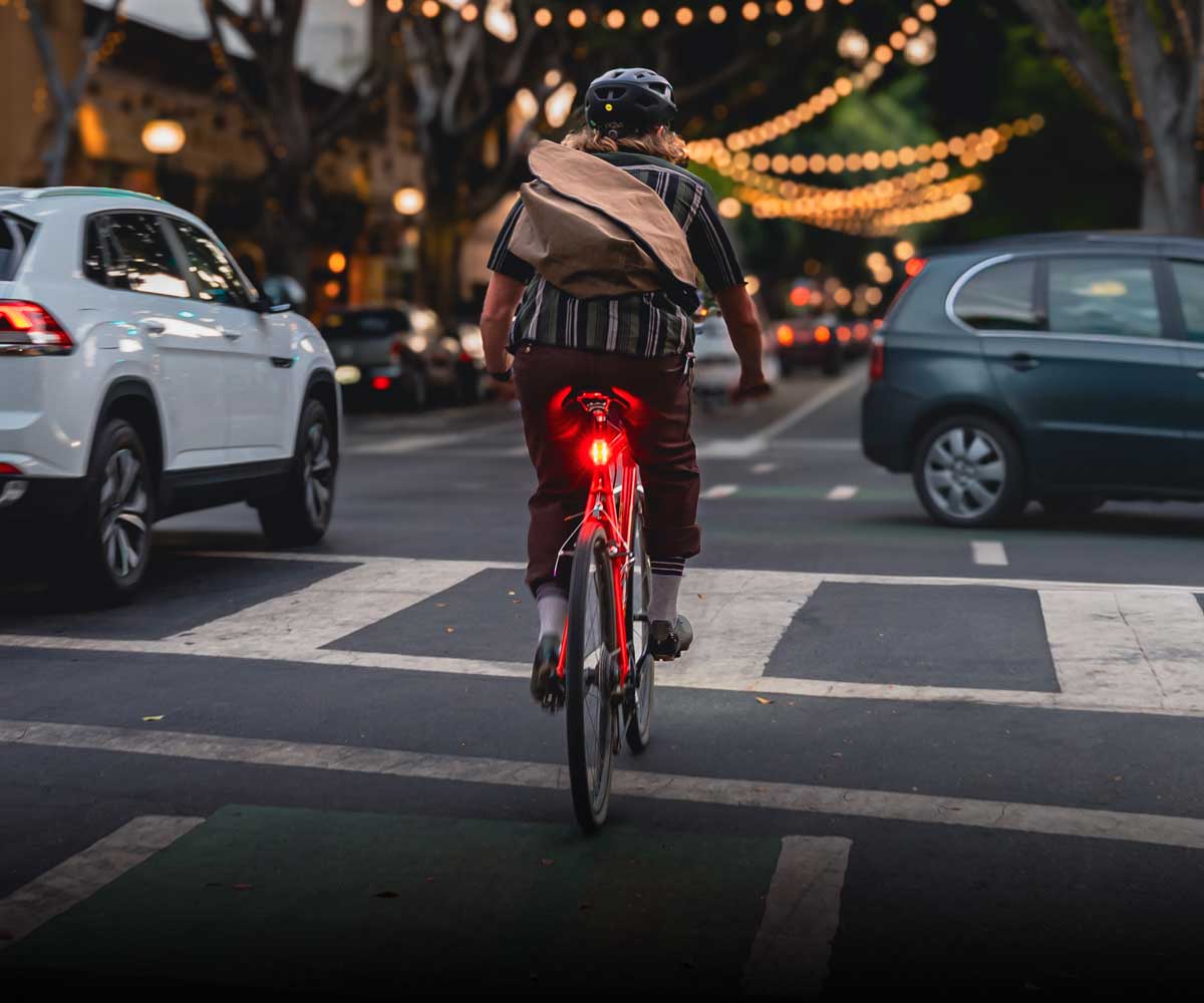 A person riding a red bicycle on a city street at dusk, viewed from behind. The rider has a bright red Radar Pro 300 tail light illuminated.