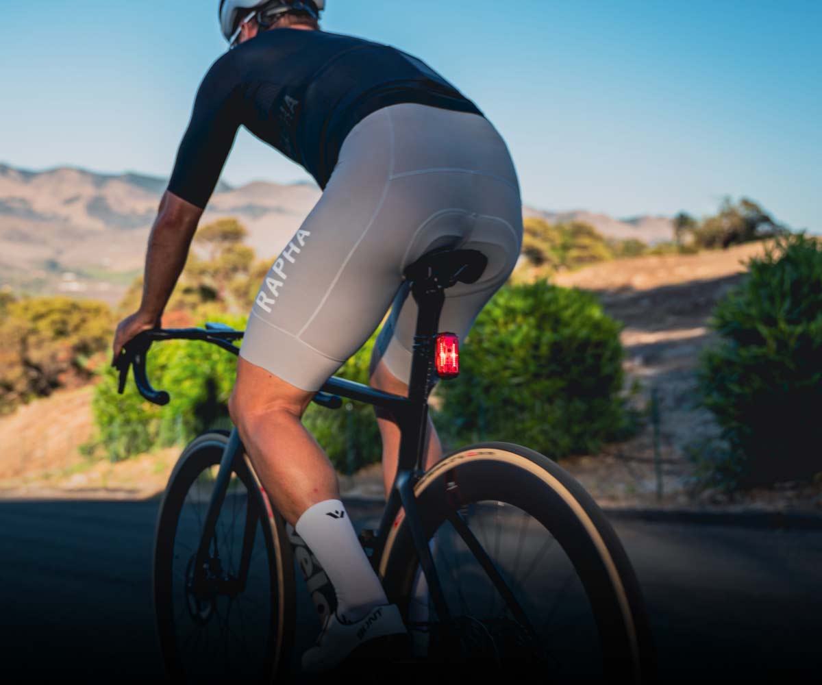 A cyclist rides a road bike on a sunny road with hills in the background while the Radar Pro 300 taillight remains bright.